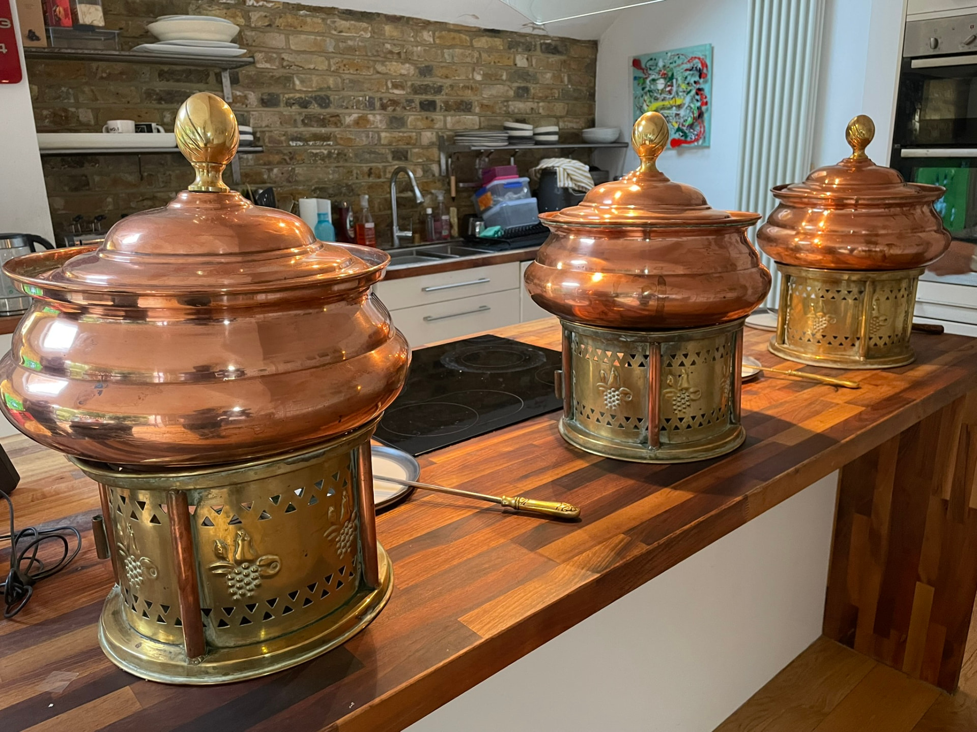 Copper and brass chafing dishes set out along a long wooden kitchen island with a serving ladle